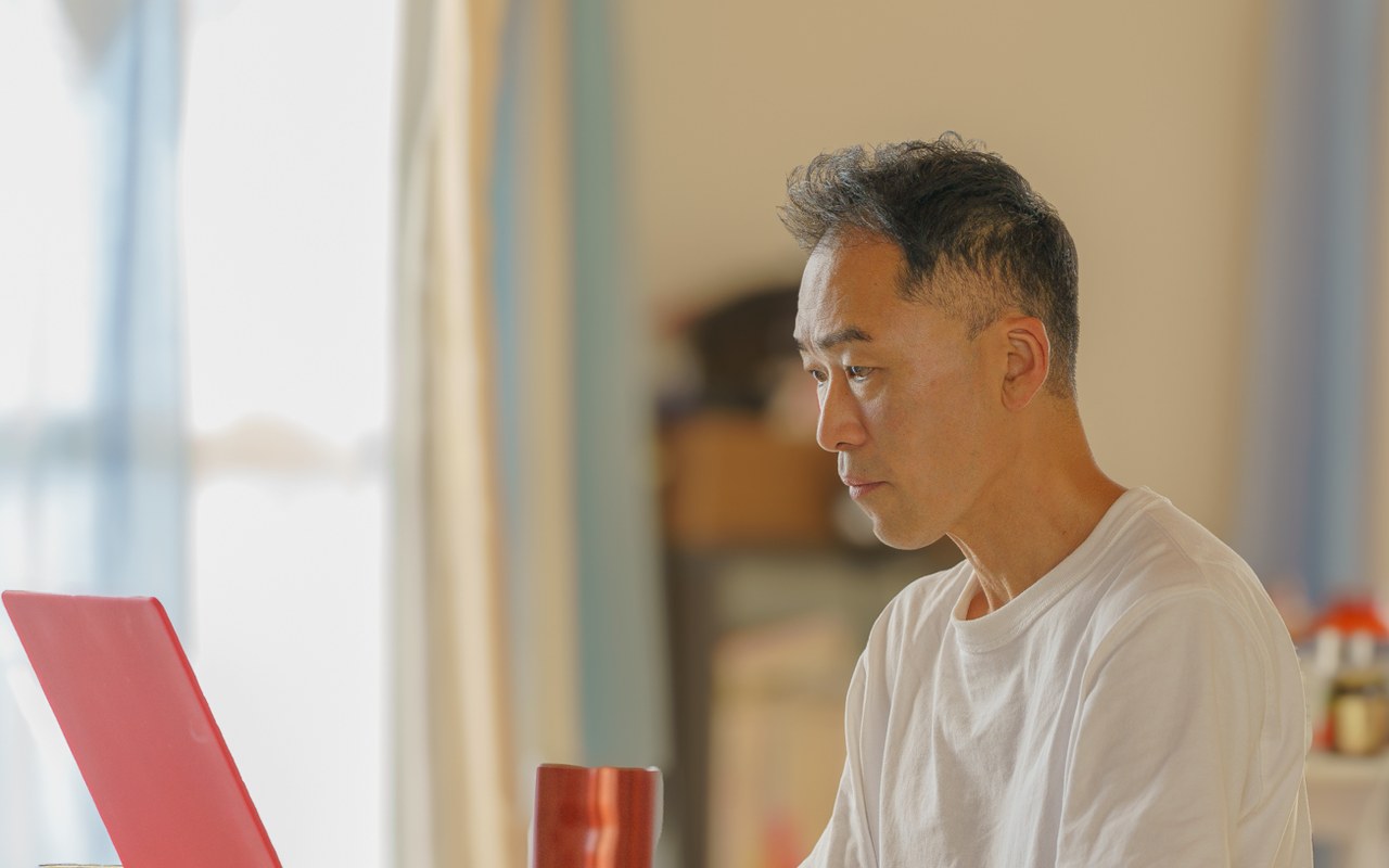 自宅の窓辺で、赤いノートパソコンに向かうRyujiさん