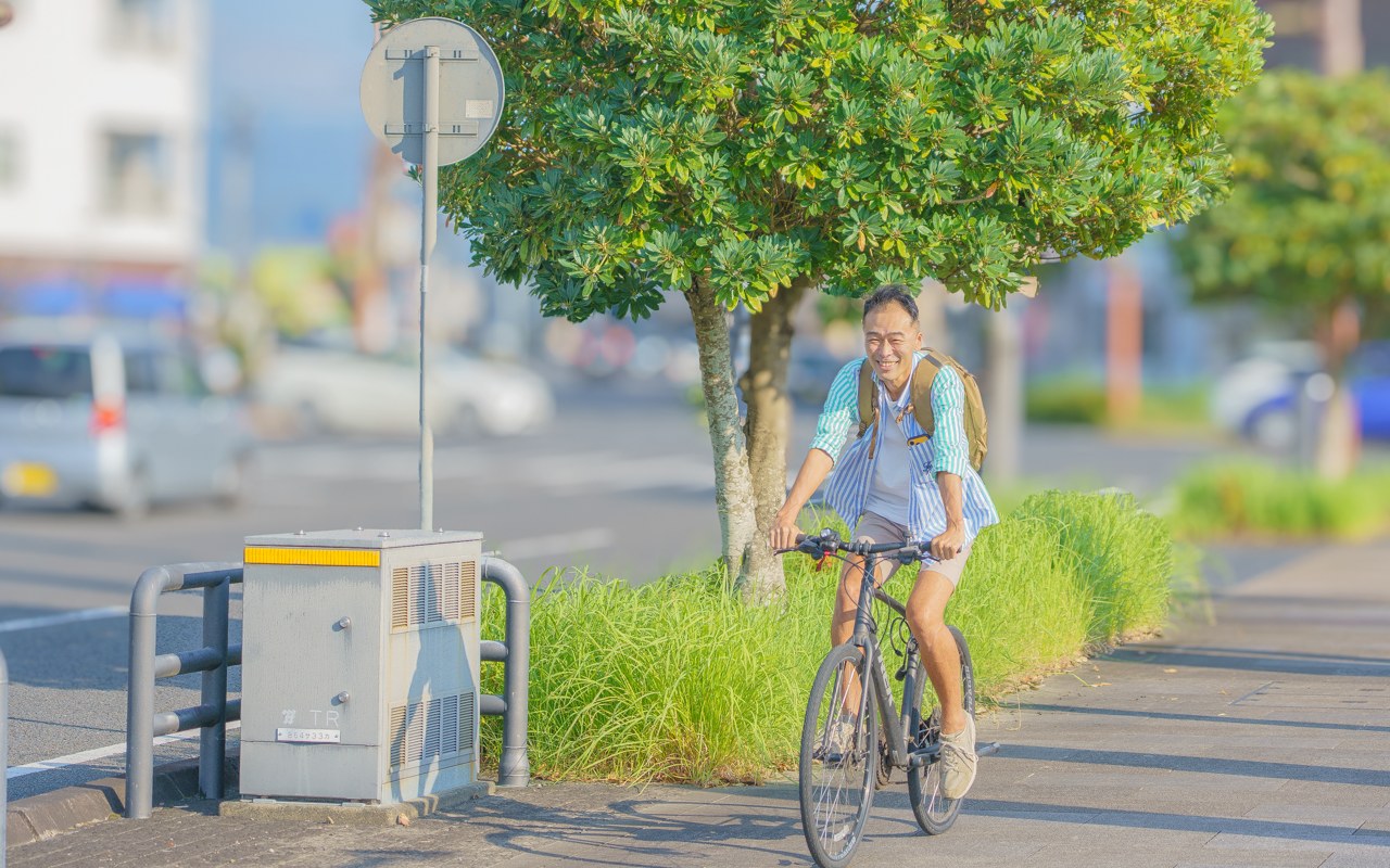 木々のある街の歩道を、自転車で移動するRyujiさん